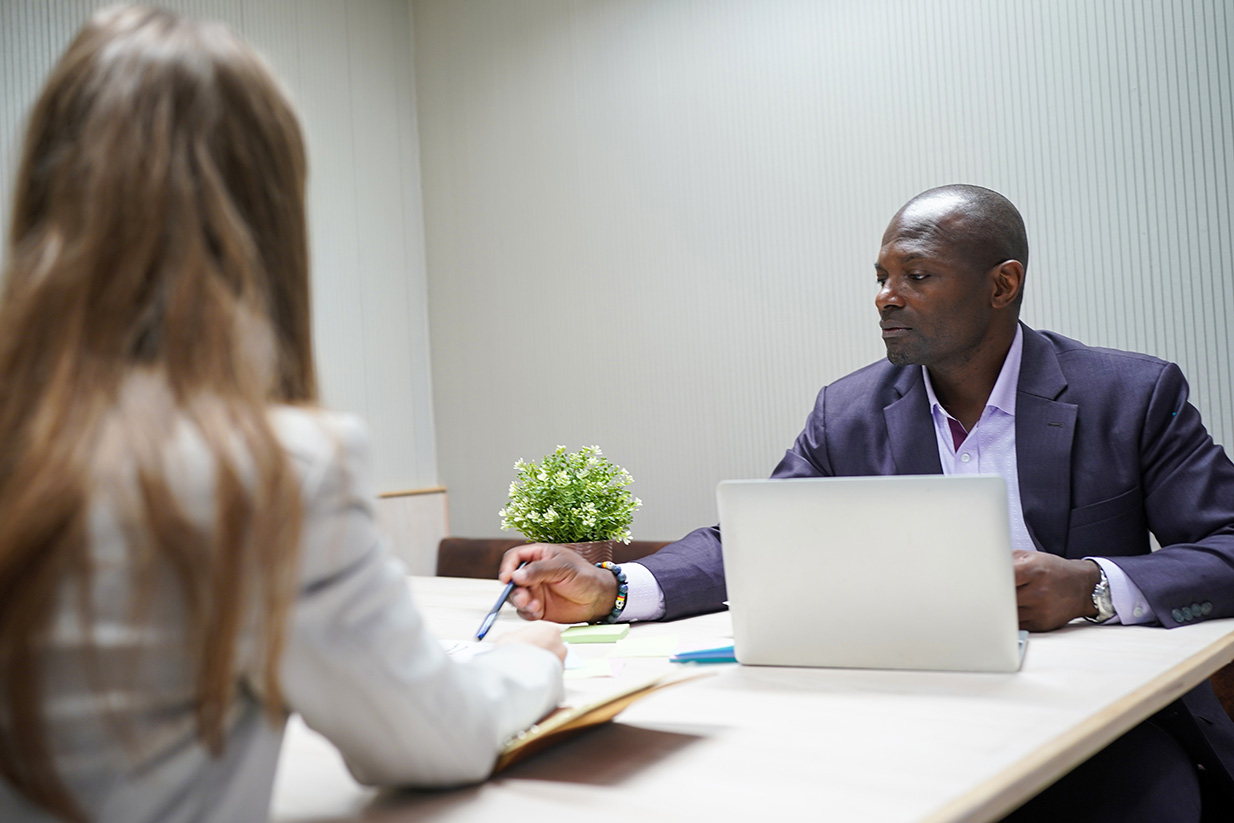 Two professionals sit across from each other at a light wood table in a meeting room; laptop open on the man's side in a purple-blazer.